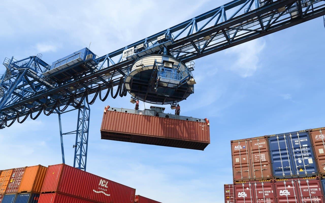 Ship-to-shore gantry crane lowering a shipping container over stacked boxes at a port — container handling and freight forwarding training